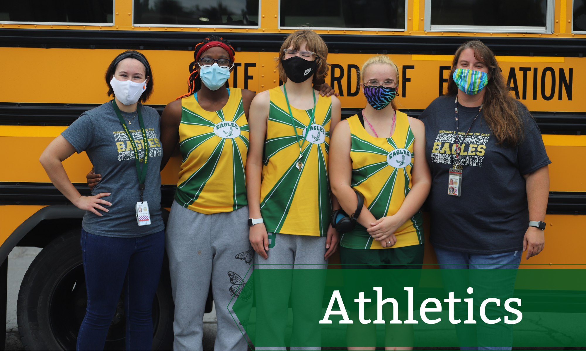 Three MSD Cross Country students and their coaches posing for a picture in-front of an MSD bus.