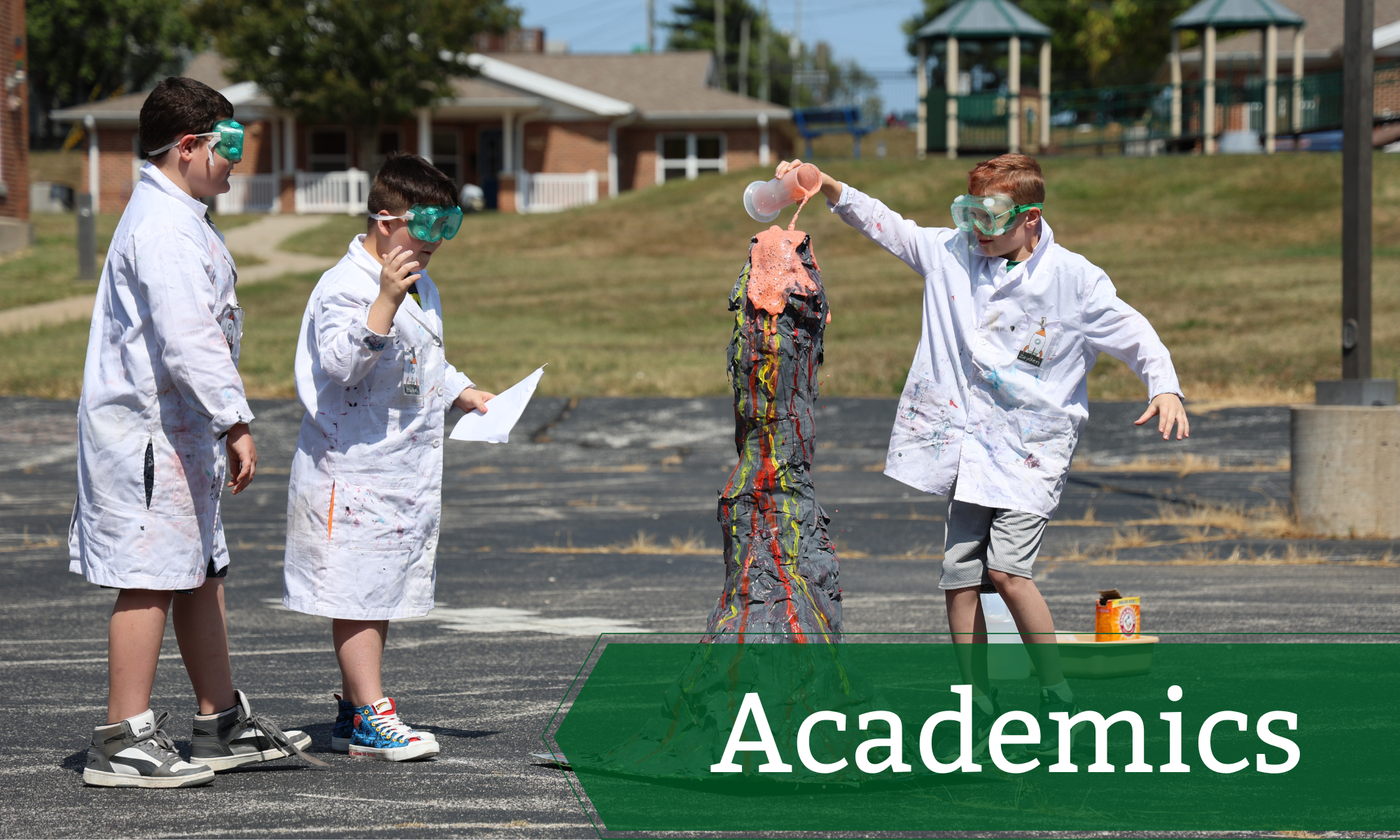 Photo of three students outdoors with a three foot paper mache volcano. The students are wearing lab coats and goggles while pouring liquid into the volcano to trigger an eruption.