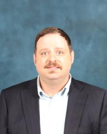 White man with brown hair and mustache wearing a dark suit jacket and light grey dress shirt.