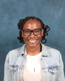 Black female with black hair in braids wearing glasses, a white shirt, and jean jacket.