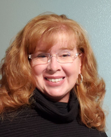 Photo of a white female with red hair and glasses, wearing a black turtle neck shirt.